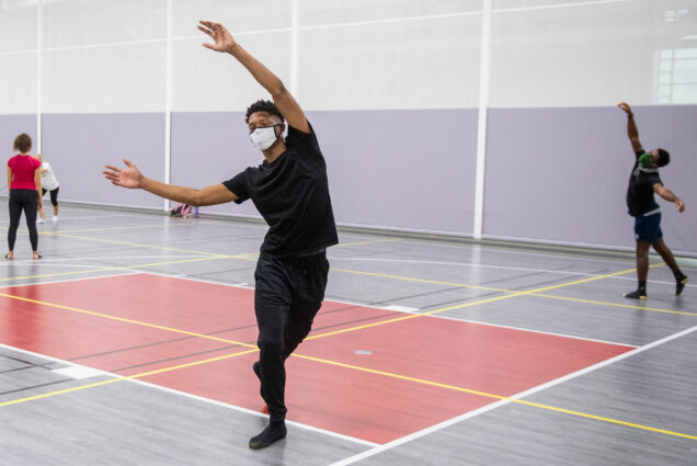 Photo of Bishop Edwards (CFA’23) moving within his 14-foot square during instructor Liz Roncka’s improv dance class at FitRec September 3. The yellow tape grid is there to ensure physical distancing. Bishop raises both his hands in the air gracefully and wears a blue mask.