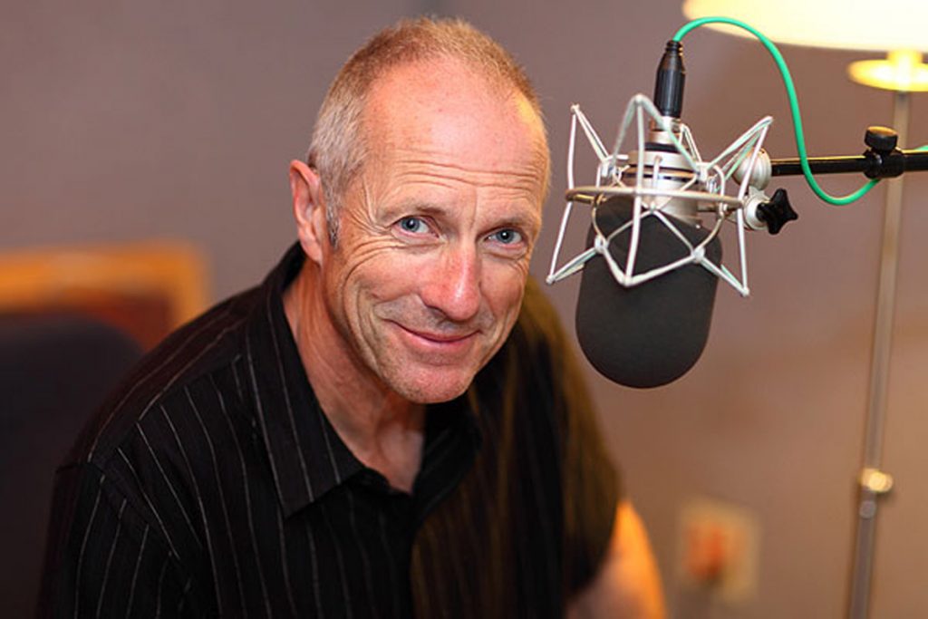 Portrait of Will Lyman at a studio mic. He wears a black t shirt and smiles.