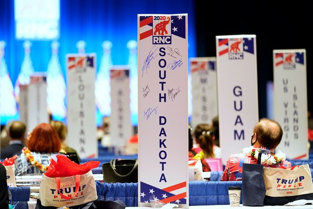 Photo from Monday, Aug. 24, 2020, as delegates watch as the roll call vote of states continues after Vice President Mike Pence spoke at the 2020 Republican National Convention in Charlotte. In the photo, large white posts with RNC branding with state names are seen, such as South Dakota, Guam, and US Virgin Islands. The back of a few people's heads are seen in the background.