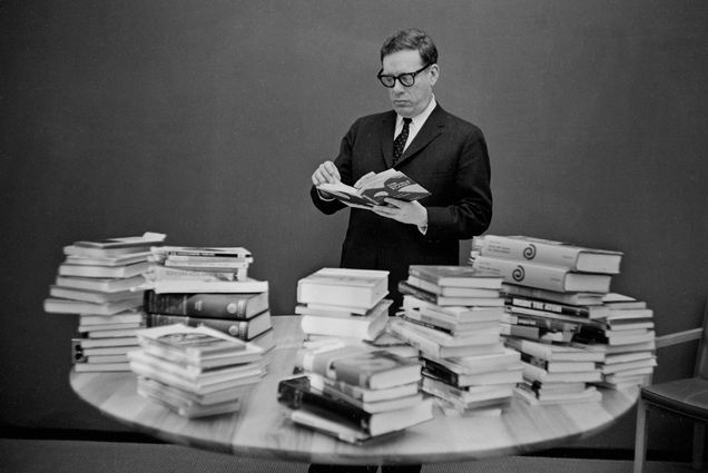 A black and white photo of author Isaac Asimov with 80 books at a Mugar Library event comemmorating the publishing of his 80th book in 1967. The table in front of him is piled high with books; he flips through one, wearing dark thick-rimmed glasses and a suit.