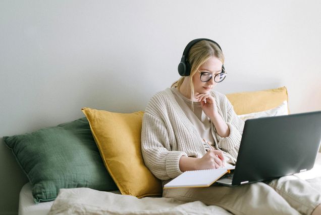 Photo of a young woman in a sweater and headphones writing notes as she looks at her laptop.