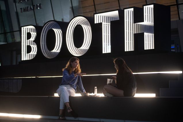 Photo of Emma Weller (CFA’24), left, and Isabella Lampson (CFA’21) chat on the steps of Booth Theater September 16. The bright white letters of the 'BOOTH' sign are seen behind them, it's dark out.