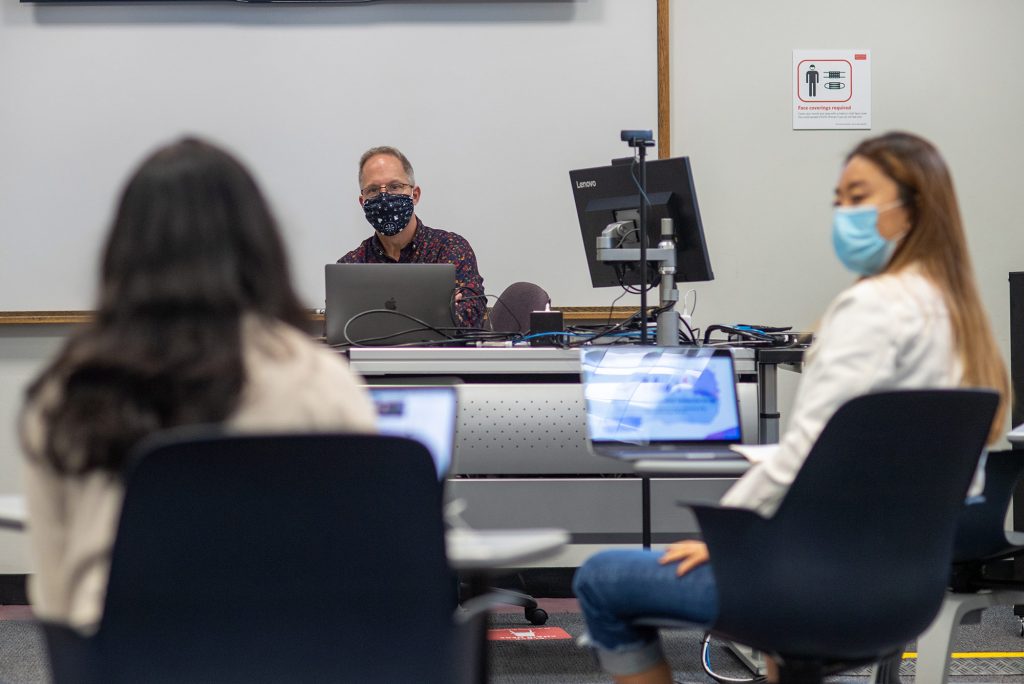 Photo of Doug Gould, a COM professor of the practice of advertising, with a mask on as he teaches in front of a computer with two students in his classroom seen blurred in the foreground.