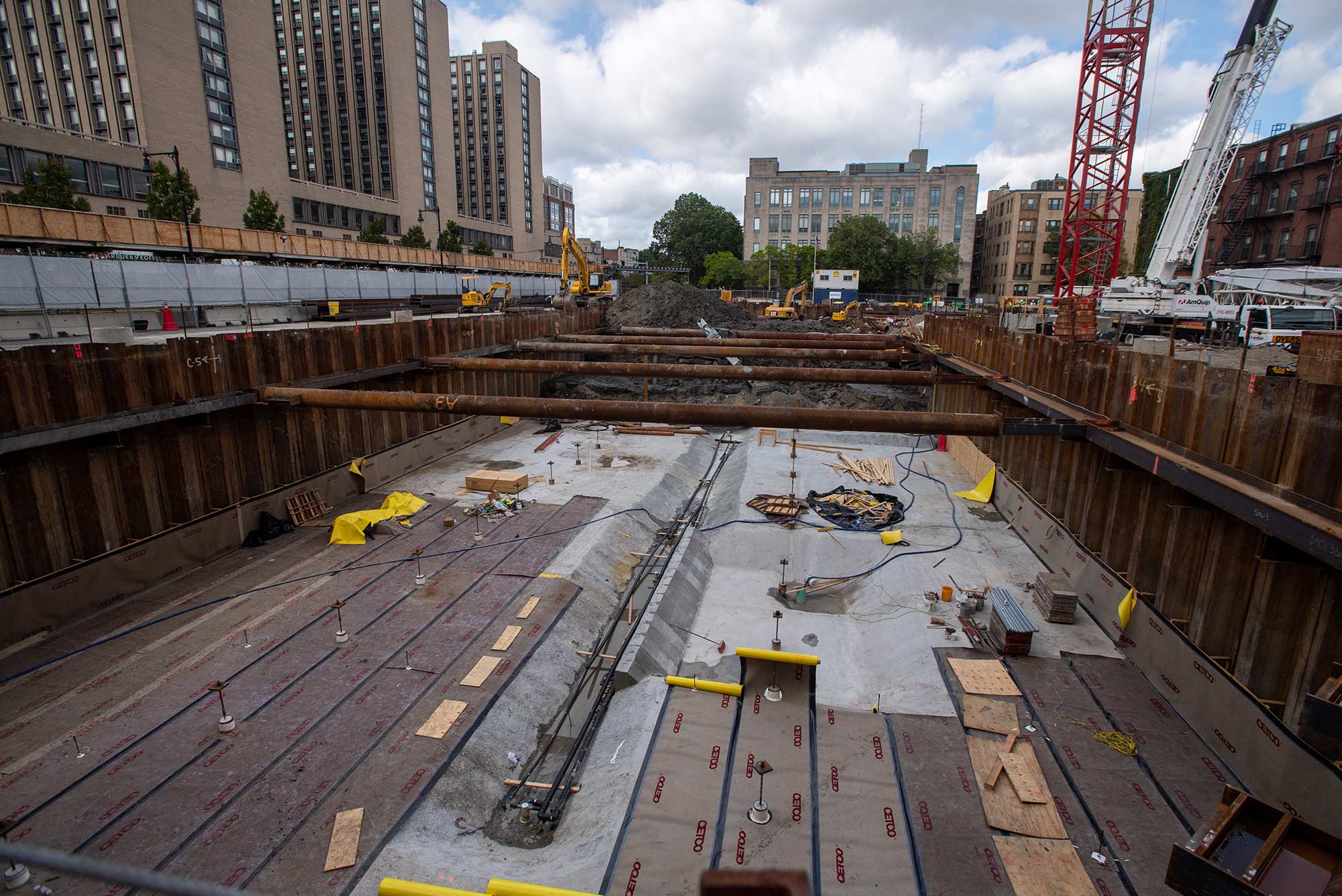 Photo of continued construction at Data Services Center. In the photo, dark colored horizontal pipes are seen. Trucks and the bottom of a red crane are seen on the right background.