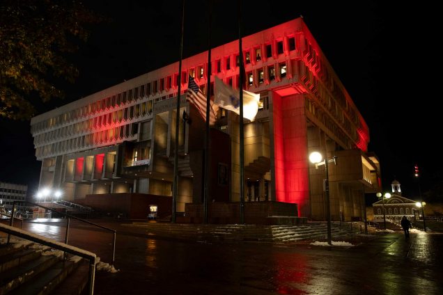 Boston City Hall lit up in red with flags flying at half mast in honor of Travis Roy