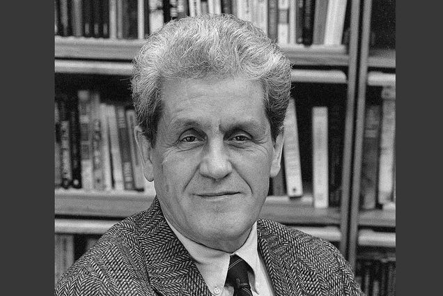 Black and white portrait of Joachim Maître in a checkered suit in front of a bookshelf.