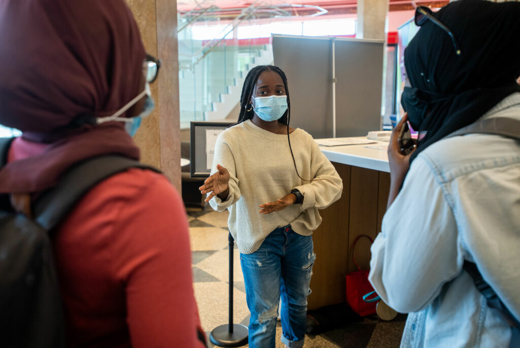 Photo of Archelle Thelemaque (COM’21) dressed in a white sweater and jeans and wearing a blue face talking to two fellow students in the Thurman Center. 