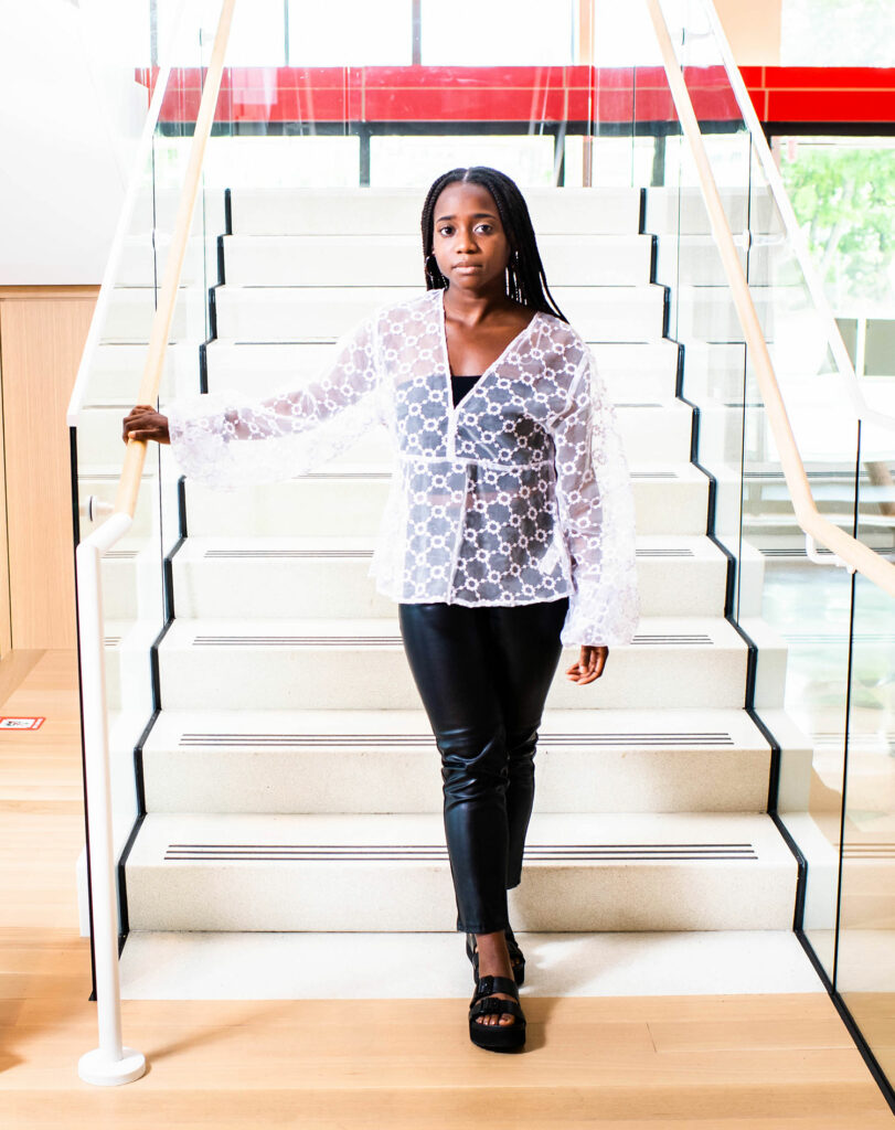 Portrait of Archelle Thelemaque (COM’21) in platform sandals and a white blouse standing at the bottom of a stairwell with her hand on the railing.