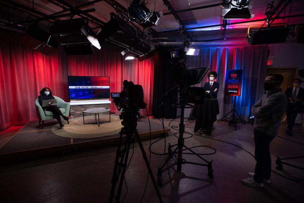 Photo of BUTV10 runs election night dress rehearsals in Studio West October 30. All the students are masked, one student sits in a chair near a tv screen, another looks at a screen behind a camera, and another host stands in the background in the dimly lit studio.