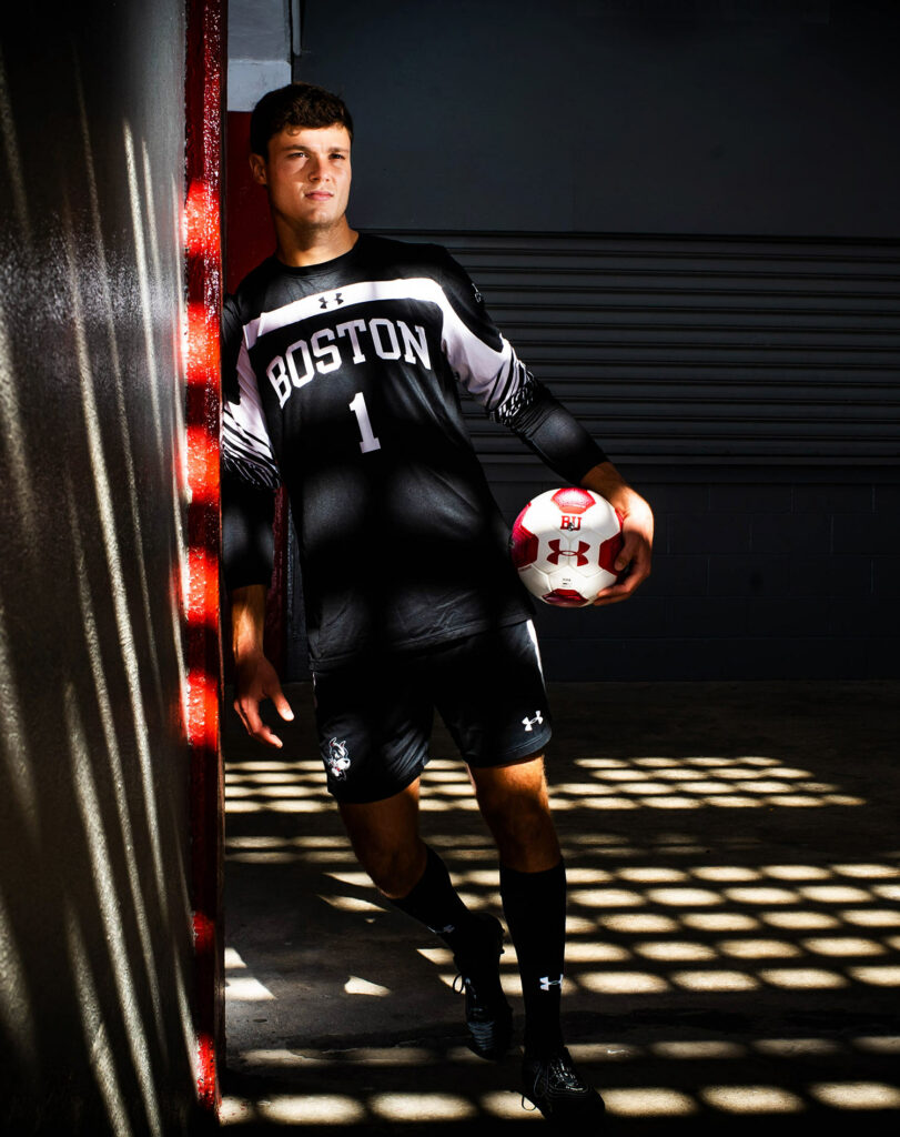Photo of Michael Stone (CAS’21)in a dimly lit locker room with light coming through in a square grid. He wears a black BU jersey with the number one and holds a red and white soccer ball to his side. 