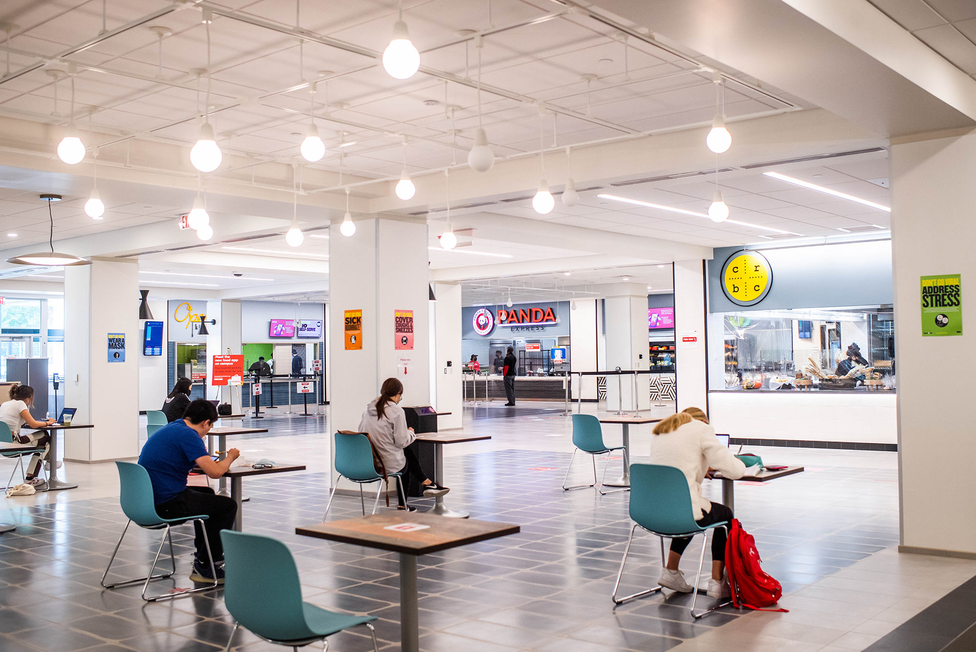 Photo du Food Hall de l'UGS récemment rénové. Des tables individuelles avec des sièges sarcelles sont étalées au premier plan, quelques étudiants s'assoient et mangent. À l'arrière-plan, on aperçoit les lumières et les murs blancs et quelques étals de nourriture.