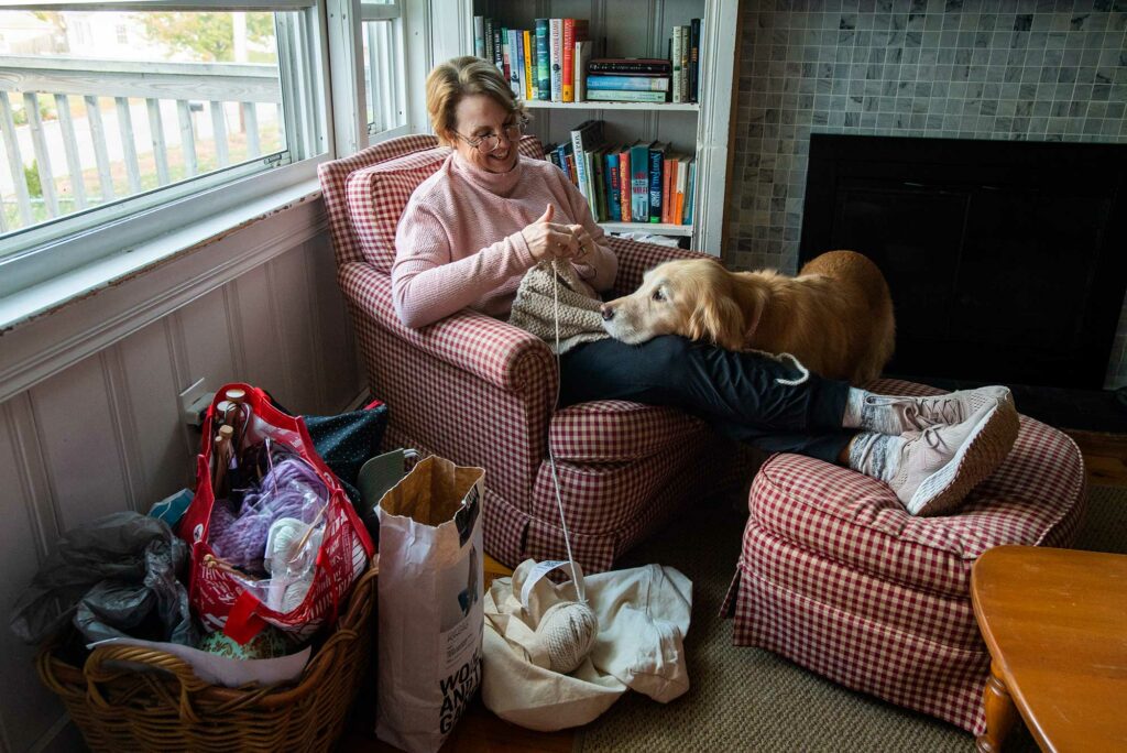 Photo of Megan Nocivelli, lecturer, administrative sciences, Metropolitan College, sitting in her home smiling with her dog on her lap as she knits. Yarn is seen in bags on the ground around her chair.