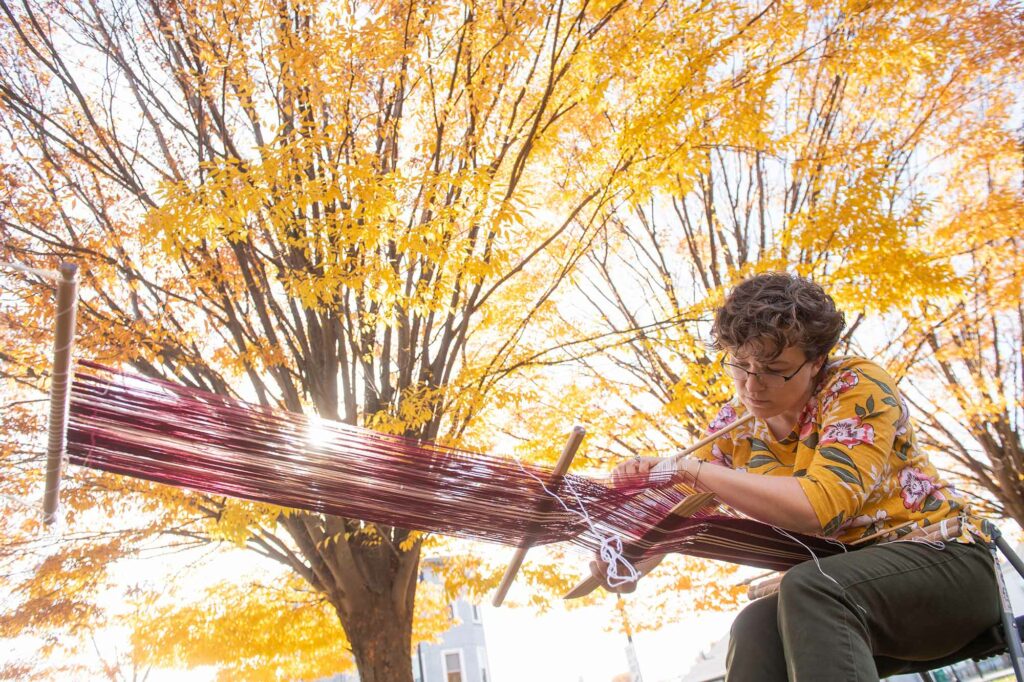 Photo of Megan Berkowitz (STH’21, SSW’21) leaning over a loom while backstrap weaving by hand. A tree with bright yellow leaves is seen in the background.