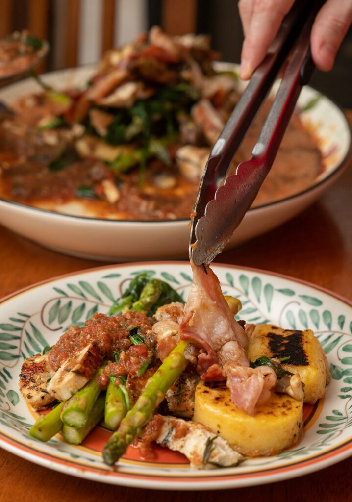 Photo of a pair of tongs plating the final piece of a dish of Jamie Oliver’s recipe for rosemary chicken with grilled polenta and asparagus in a porcini mushroom ragu sauce. A full plate is seen in the background.