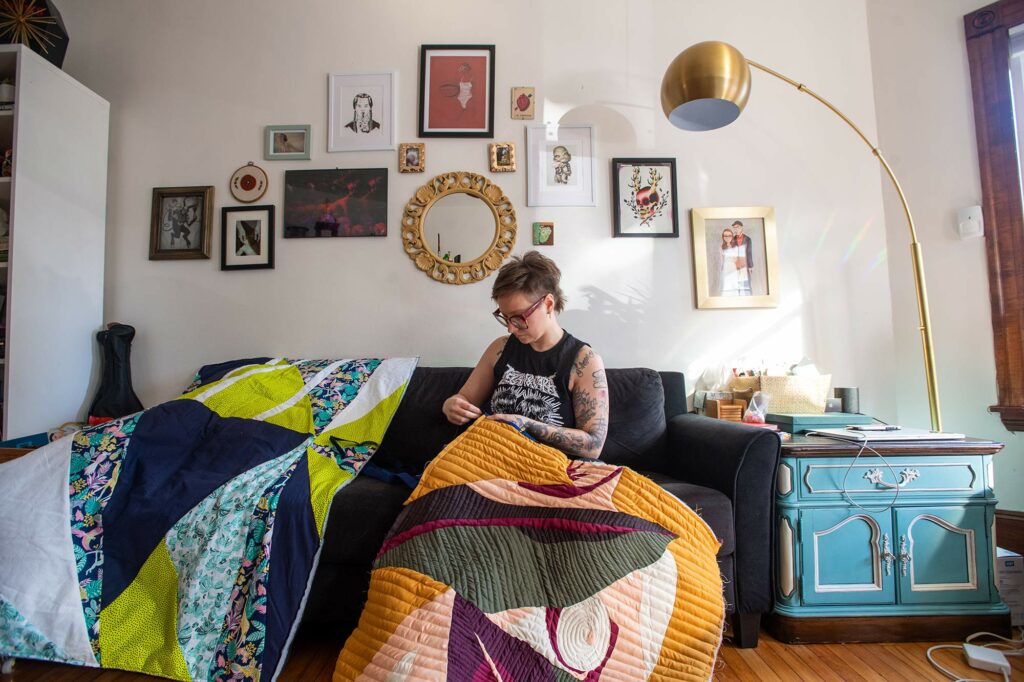 Photo of Janice Checchio, associate creative director, BU Photography sitting on her couch as she works on a large quilt with brightly-colored geometric shapes.