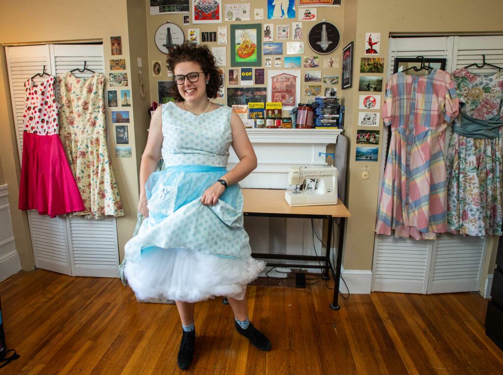 Photo of Grace Saathoff, here in her Brookline home, sewed a wardrobe of vintage-inspired dresses over the summer. Saathoff wears a light blue polka-dotted dress and smiles; other dresses she's made hang in the background.