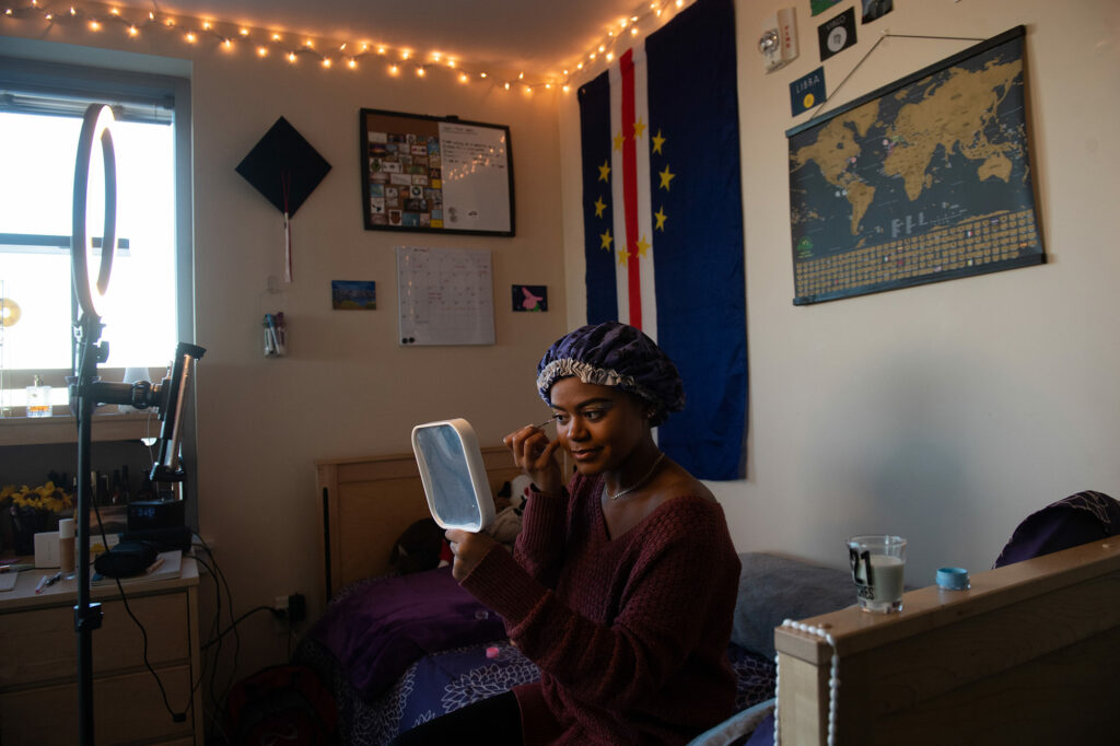 Photo of Tatyana F. Da Rosa (CAS’20, Questrom’21) smiling and sitting on her bed in a bonnet while doing her makeup. Da Rosa's Hobby is makeup and makeup tutorials.