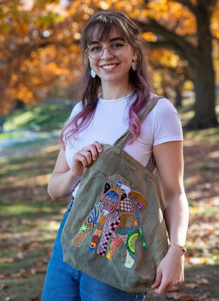 Photo of Sarah Kula (LAW’21) show off a bag she embroidered. The green bag has a brightly-colored stitches that form the shape of an elephant.