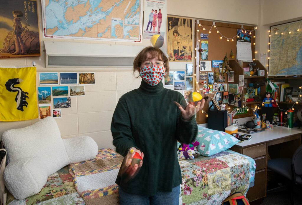 Emma French in her Warren Towers dorm room practicing her juggling. She wears a spotted face mask; her dorm walls are covered with maps, photos, and cork boards.