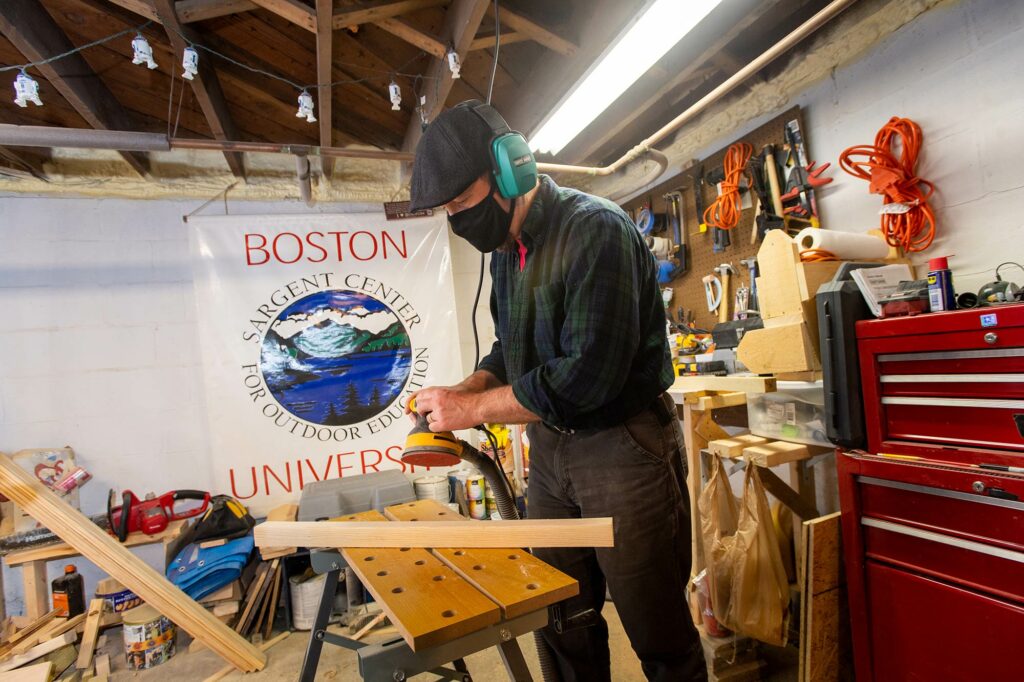 Photo of Paul Hutchinson working on a guitar stool in his woodshop at his home in Jaffrey, N.H. He wears a face mask and noise cancelling earmuffs. A large Boston University Sargent College sign hangs on one wall of his shop; the other wall is filled with tools.