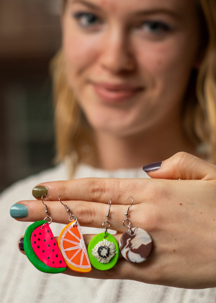 Grace shows off some of her playful earrings, a watermelon, orange, kiwi, earrings hang from her hand.