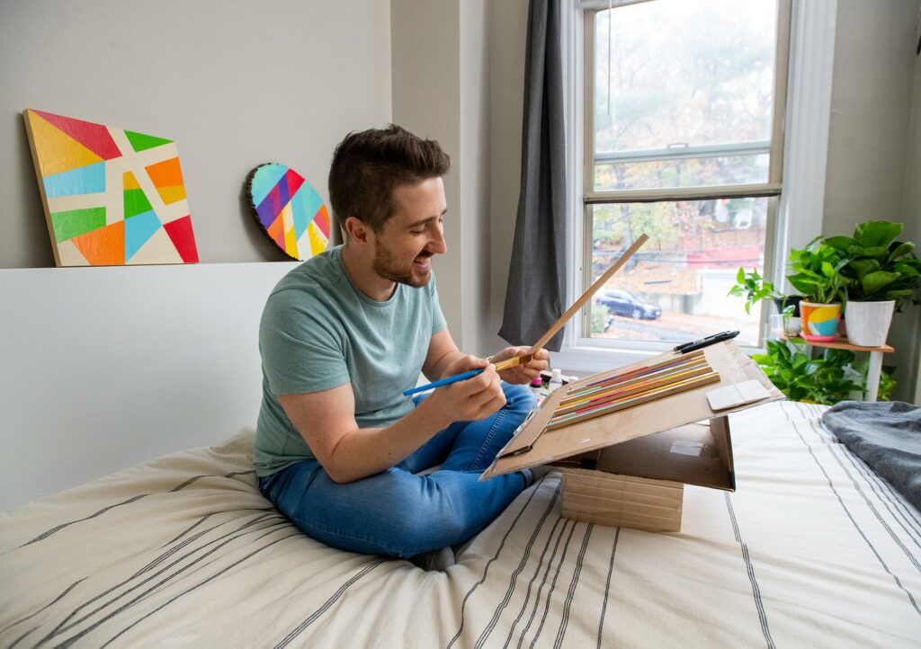 Photo of RJ Foley IV sitting on his bed working on his colorful wood art paintings in his Brighton home, with completed pieces behind him. Bright geometric shapes are seen on a circular and square piece of wood.