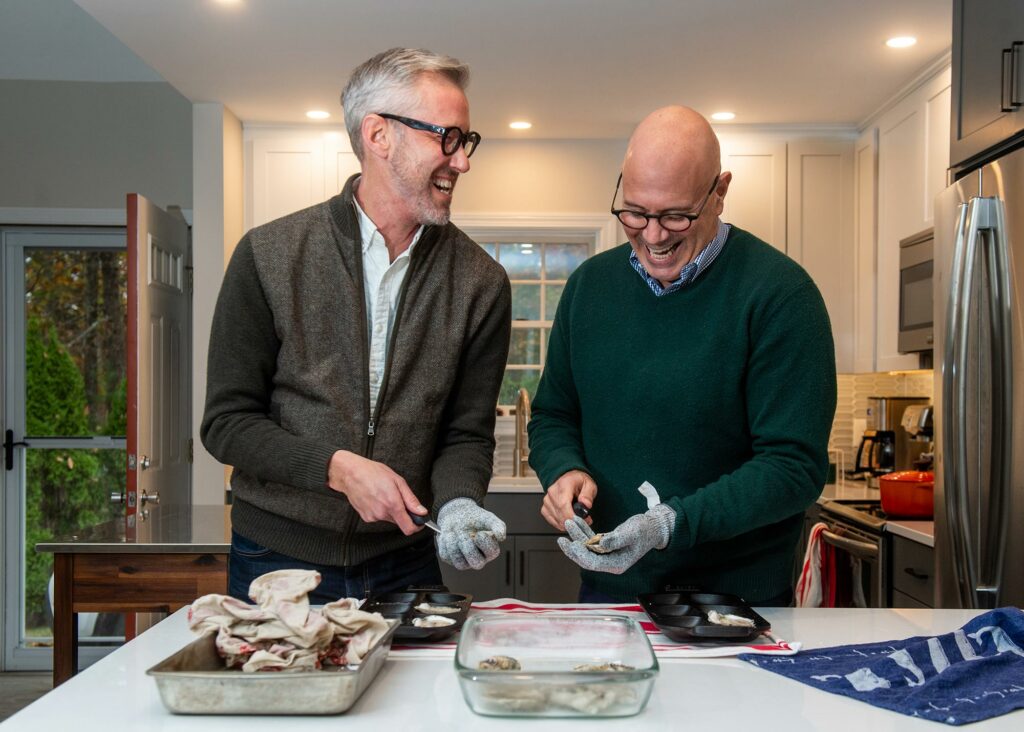 Photo of Daniel Spiess (left), with husband Marc Scatamacchia shucking oysters at their home in Ogunquit, Maine. The two smile widely and wear gloves while holding oysters; a tray of shucked and unshucked oysters sit on counter before them.