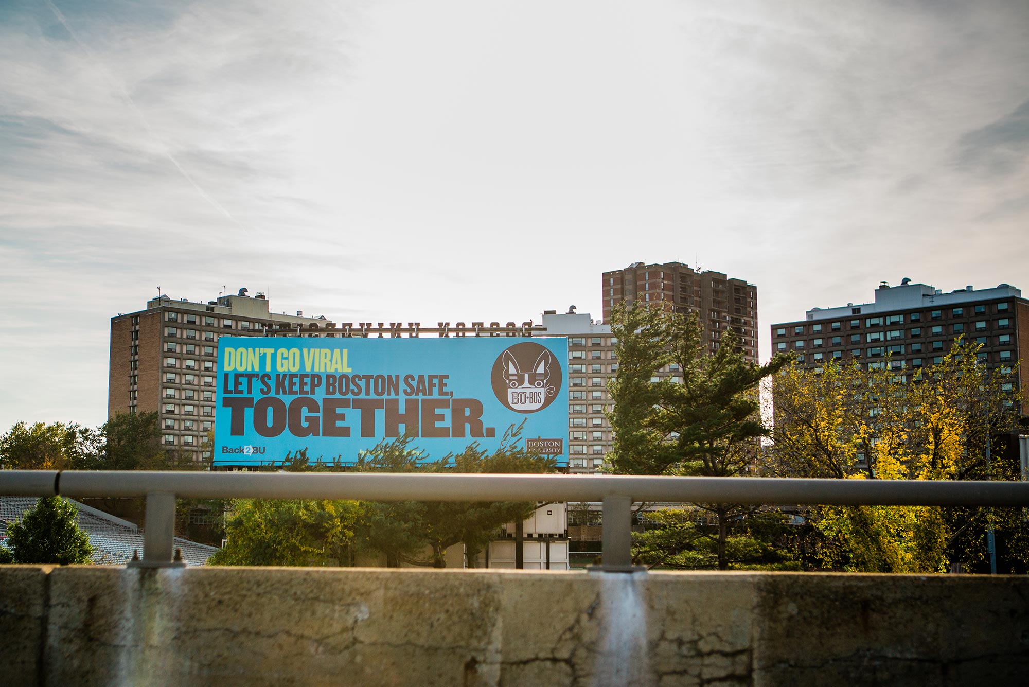 Photo of Billboard facing the Mass Pike over Nickerson Field. The sign is blue and reads "Let's Keep Boston Safe, Together." A gray cement railing is seen in the foreground, the sun sets behind the building.