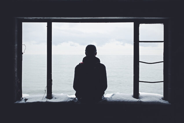 Photo of a person sitting on a snow bench under and overhang looking out to the ocean with their back to the camera. The inside of the structure is black and in silhouette, the ocean and sky are very gray.