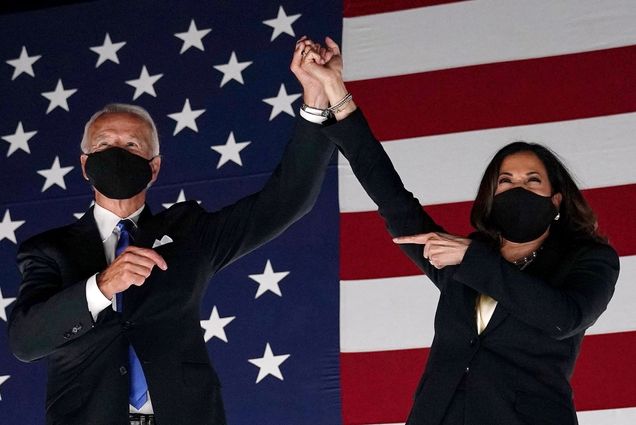 A photo of president-elect Joe Biden and Vice President-elect Kamala Harris clasping hands and raising them above their heads. They are standing in front of an American flag. Both are wearing masks.