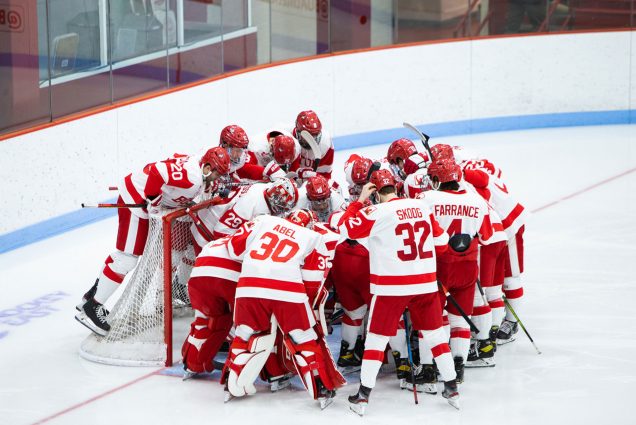 A photo of the BU men's ice hockey team huddles up on the ice around one of the goals