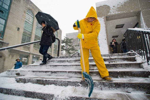 Photo of Jose Ferreira in a yellow rain suit clearing the snow from the steps near Mugar Memorial Library in 2011 as a student with a black umbrella walks by. Snow falls around him.