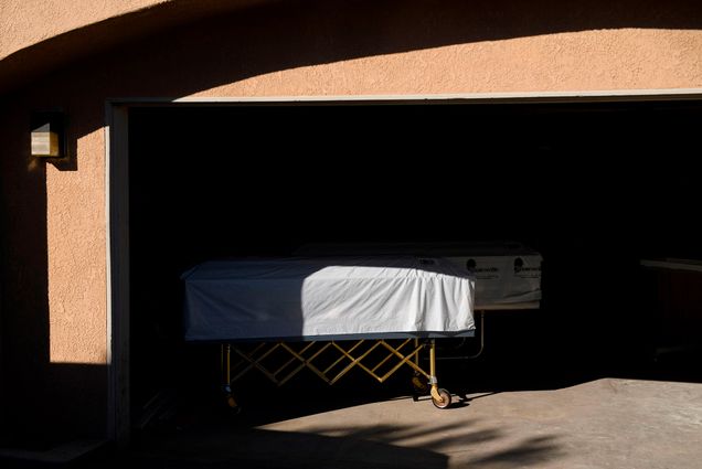 Photo of a casket covered with a white sheet over it on a dolly at the edge of a garage; it is partly in the shadows and the garage is darkened.