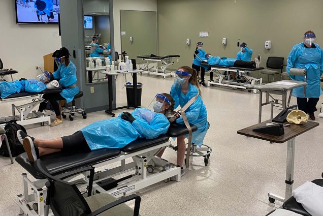 Photo of students in Sargent’s occupational therapy program in full blue PPE gowns, face shields and masks, during a training session. One student lays on the bed while the other attends to mock-patient.