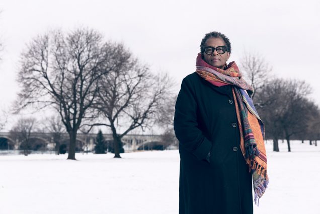 Photo of Henriquez, a middle-aged Black woman, standing in a long p-coat and colorful scarf in a snowy park.