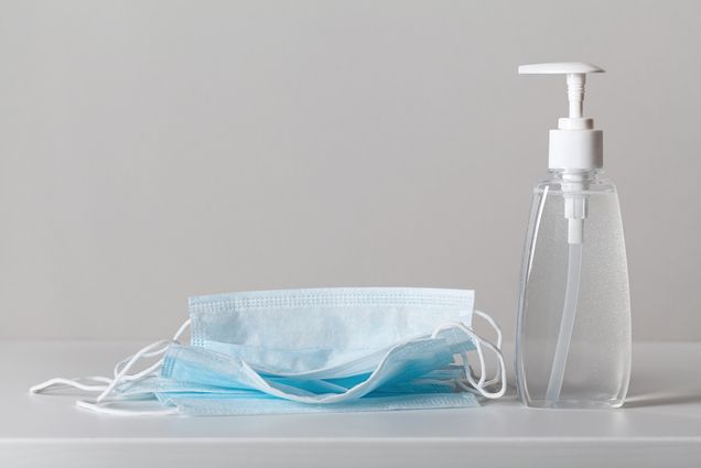 Photo of a pile of blue medical face masks on the left and a bottle of hand sanitizer on the right; they both rest on a white surface.