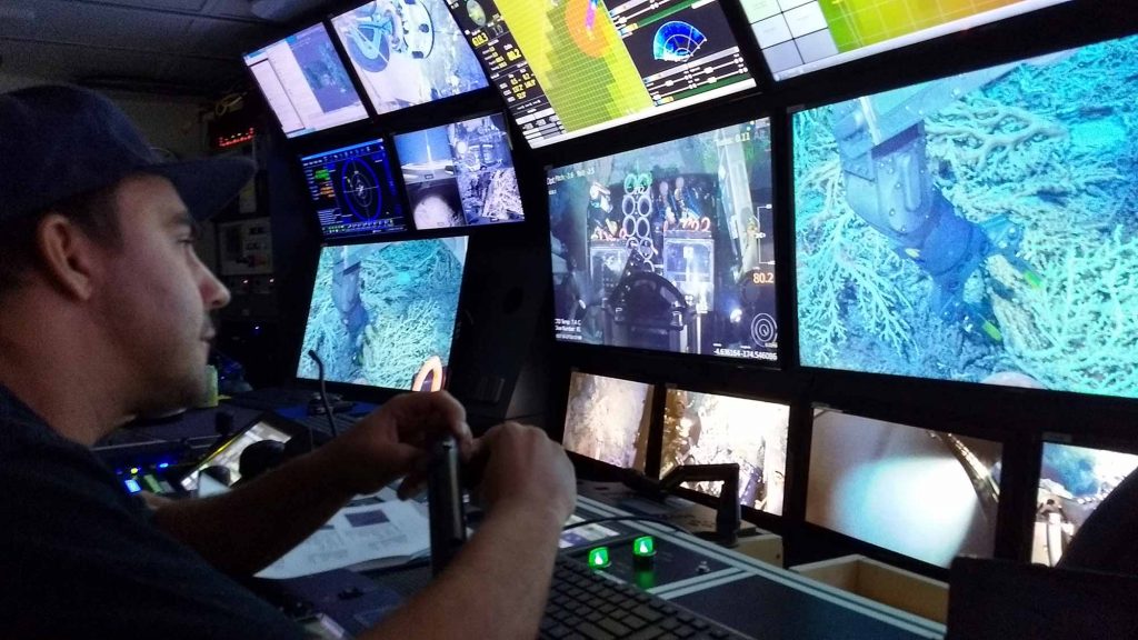 ROV pilot Adam Whetmore flies the submersible SuBastian in front of a wall of screens in a cockpit onboard the R/V Falkor .