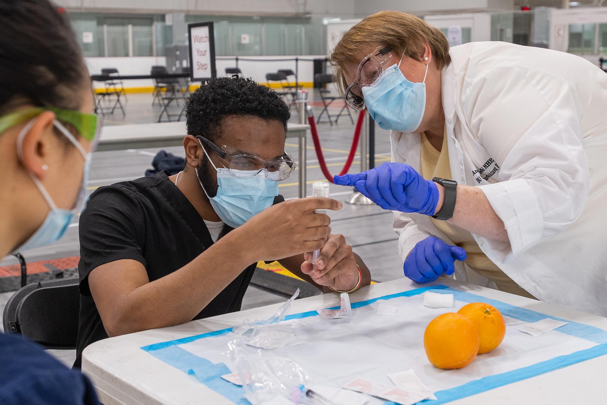 Photo of Rose Zhao (MED’24),left, who looks on while director of clinical education for the BU PA program Angela Reffel, in a white coat and blue gloves, advises Micaiah Bully (MED’24) at the vaccination administering training at FitRec March. Bully looks intently at a vial and syringe. Oranges rest on the table.