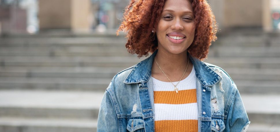 Photo of Nyah Jordan (CGS’20, COM’22) standing in front of the steps in Marsh Plaza. She smiles, has curly shoulder-length reddish hair, and wears a jean packet and gold and white striped sweater.