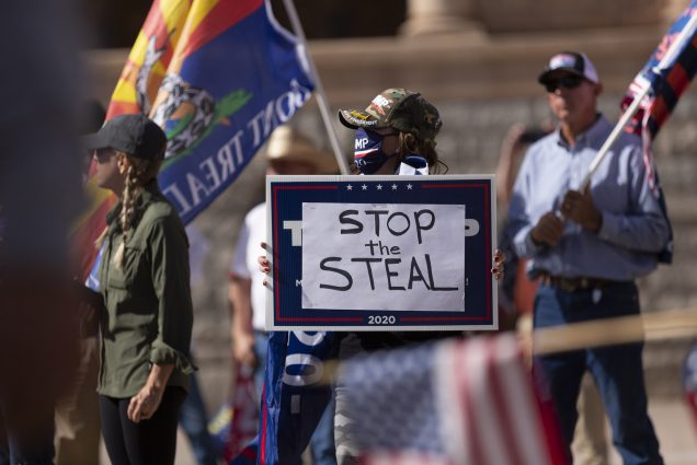 Photo of a ‘Stop the Steal’ in early November 2020. A group of people stand with ‘don’t tread on me’ flags and American flags. In center foreground, a person in a camo baseball cap holds a sign with the words ‘Stop the Steal’.