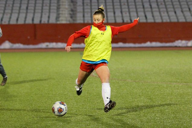 Photo of Ann Marie Jaworski (CAS’20, Wheelock’21) in a red long sleeve, shorts, and face mask, with a bright yellow practice pinny, about to strike a soccer ball. Empty bleachers and the foot of a team mate are seen in the background.