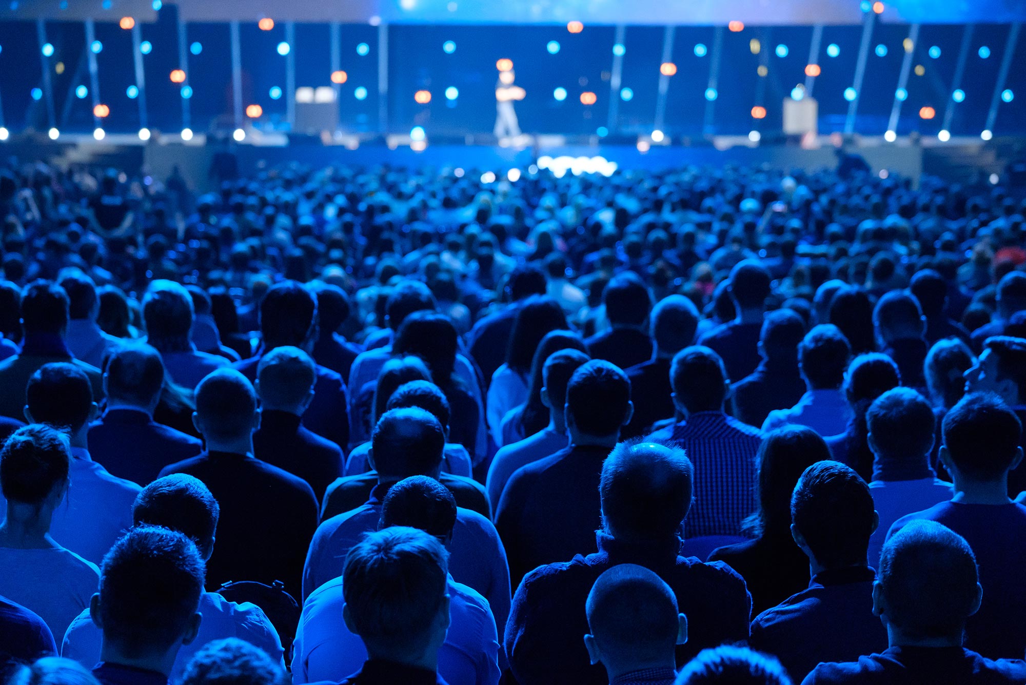 Photo of a large performance/event venue filled with people who listen to a speaker on stage. The photo is taken from the behind the audience and captures the blue-lighting used at the event.