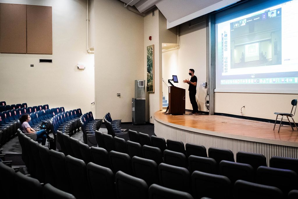 Photo of Samantha Haas (COM’22) (left) is the lone student in attendance during College of Communication lecturer Mike Proulx’s advertising class on September 14, 2020. Proulx stands behind a podium on the stage with Zoom projected behind him.