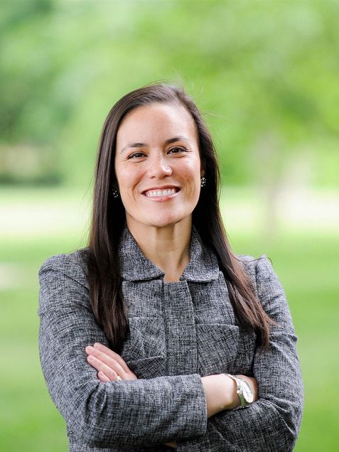 Photo of Gina Ortiz in a gray blazer with her arms crossed, smiling, with green space blurred behind her.