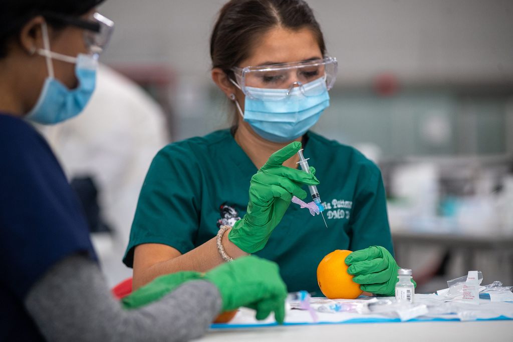 Photo of Tia Patterson (DMD’24) practicing administering a vaccine on an orange, she raises a syringe above the orange. She wears goggles, a blue face mask, a green shirt and green gloves.