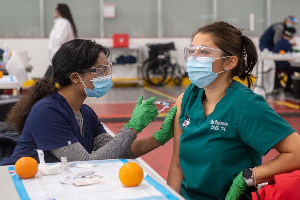 Photo of Second year BU med student Alekha Kolli administering a practice vaccination of a simple saline solution on Tia Patterson (DMD’24) at the vaccination administering training at FitRec. Both wear face masks and goggles, an orange rests on the table in front of them.