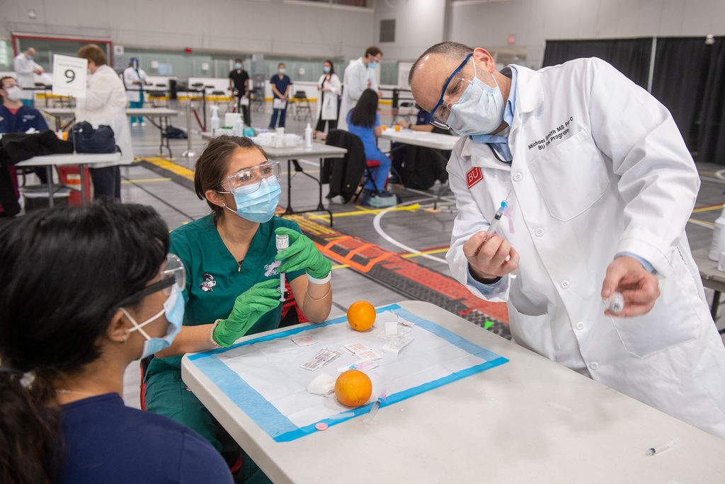 Photo of BU med student Alekha Kolli, from left, and Tia Patterson (DMD’24) learning  from Michael Smith, MS PA-C at the vaccination administering training at FitRec March 7. They all wear face masks and goggles and sit around a table in the middle of the gym.