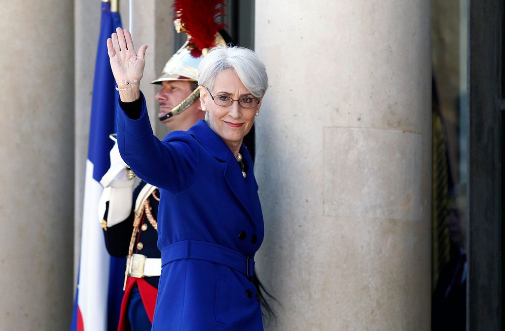 Photo of Wendy Sherman, in a blue blazer, waving her hand. Behind her, a soldier holding a flag is seen.