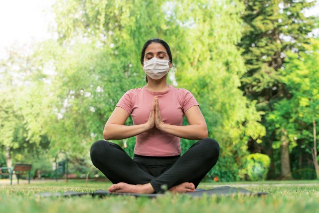 Photo of a woman in a pink shirt, face mask, and black leggings doing yoga outside in a park with lots of greenery.