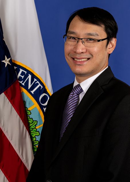 Headshot of Nicholas Lee in a purple tie with a black suit jacket on in front of an American and DOE flag.